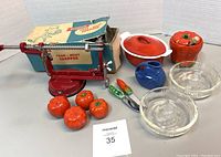 Full view of the vintage kitchen lot including red food chopper, cast iron cassoulet, tomato-themed ceramics, apple peeler, glass juice reamers, and pea pod-shaped utensils.