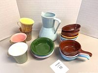 Photo showing three rattan-style plastic mugs with yellow lids on the left, a light blue Brush McCoy ceramic pitcher in the center, and several stacked ceramic dishes in brown, orange, blue, and green colors on right.