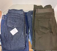 Lot of five pairs of ladies pants, blue denim and olive green colors, folded and stacked.