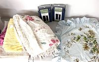 View of assorted linens including chenille bedspread, pillow shams with floral embroidery, yellow cloth, and two packaged navy drapery panels.