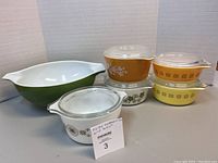 Photo shows green Pyrex Cinderella bowl alongside five small Pyrex casseroles with glass lids, displaying varied floral and starburst patterns.