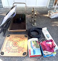Wooden decorative tray with red and black painted circular design, metal cart frame with wheels, two brass fireplace tools, coiled black hose, box labeled 'Gourmet Stix', and blue wired basket with various items on pavement