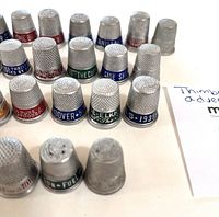 Close-up photo of multiple metal advertising thimbles lined up showing the tops and advertising bands with various brand names and slogans