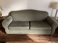 Photo of a green fabric couch with two seat cushions and rounded armrests placed against a wall with a lamp on each side.