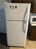 Front view of white Frigidaire refrigerator with top freezer closed, with magnets on freezer door, standing on kitchen floor near wooden cabinets and microwave.