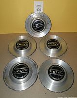 Front view of 5 Volvo metal hubcaps arranged on table showing worn silver finish and black Volvo emblems.