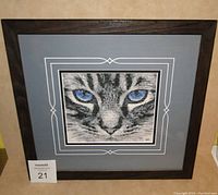 Front view of framed cross stitch depicting cat's face with blue eyes, showing detailed matted layers and frame.