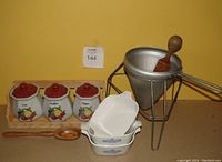 Photo showing antique wooden-handled berry masher with metal strainer on stand, 3 ceramic fruit-themed cannisters with red lids labeled Sugar, Coffee, Tea; 3 Corningware Blue Cornflower baking dishes without lids, all on plain surface with yellow background