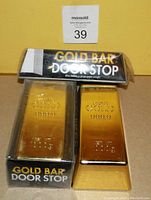 Two fake gold bar door stops with original packaging. One door stop is out of box, one inside box, both gold-colored with incised fine gold markings.