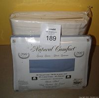 Photo showing cream color sheets still in shrink wrap and blue fitted sheet in labeled packaging with Natural Comfort branding and thread count details.