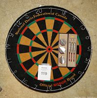 Front view of the round NODOR branded bristle dart board with accompanying three darts and additional flights in packaging, set on a tiled floor.