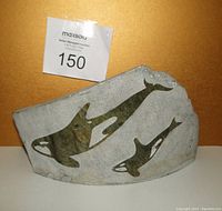 Front view of soapstone carving showing three inlaid whales on gray stone base with MaxSold auction label.