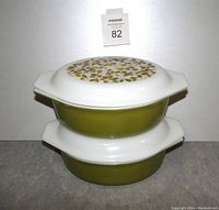 Two vintage Mid-Century Modern Pyrex casseroles stacked, showing olive green body with white lids decorated with olive pattern.