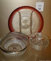 Front view of three glassware items: cranberry glass divided tray standing upright in the back, crystal bowl with silver-tone rim in front left, and lidded pressed glass candy dish in front right.