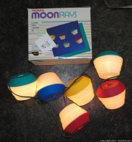 Photo showing six lit vintage Noma Moonrays patio lanterns with colored tops and the original box behind them, displaying product branding and specs.