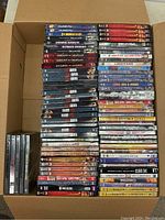 Photo showing a cardboard box filled with approximately 80 DVDs stacked in rows. Titles visible include workout programs (P90X, Insanity), several movie titles and TV series sets. Condition of cases varies, some appear new others show damaged packaging.