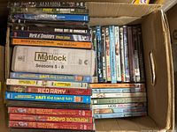 Top view of assorted DVDs and video game DVD cases in a cardboard box showing various titles and conditions.
