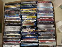 Box containing multiple DVDs stacked horizontally and vertically showing various titles and conditions, some boxes damaged or worn.