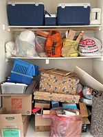 Full view of shelves with blue ice chests on top, bags of crafting supplies, blue plastic bins, folded patterned cardboard, and cardboard boxes stacked below.