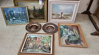 Five framed paintings and two decorative metal plates arranged on floor depicting various subjects including landscapes and cityscape