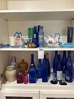 Collection of various glassware including tall cobalt blue bottles, small colored glass bottles, ceramic vases, and decorative porcelain pieces arranged on shelves.