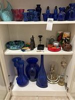Shelf with multiple cobalt blue glass vases, candleholders, decorative clock, and assorted collectibles including ceramic fish and colorful bowls.