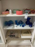 Shelf view showing several blue glass Avon items including vases and perfume bottles, some green and clear glass bowls also present, 1963 Life magazine stack at bottom shelf.
