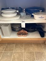 Shelves with assorted stackable white Corelle plates, glass and ceramic bakeware with blue cornflower pattern, blue covered pot, stainless steel pots, non-stick frying pans, and white Tupperware container.