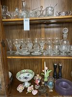 Crystal glassware collection including candlesticks, various drinking glasses, a cut-glass decanter, and a decorative crystal compote. Also includes purple glass plates, Capodimonte porcelain rose flowers and ceramic bowl, salt and pepper mills, and other decorative items.