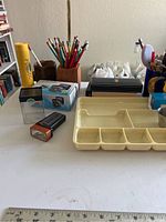 Close-up of stationary supplies on the table including pencils in holders, a yellow container, small boxes, a flashlight, and a multi-compartment plastic organizer.