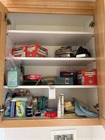 Three shelves inside a wooden cabinet containing various laundry and cleaning supplies including boxes of UltraSoft tissues, Surf soap bars, folded cloths, sponges, and small household items.