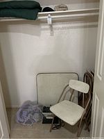 Folded vintage card table leaning against closet wall with one white folding chair in front, mesh aqua jogger visible on floor.