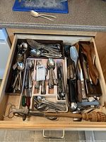Open kitchen drawer with multiple compartments containing forks, spoons, knives, and various kitchen utensils including wooden spoons and plastic handled tools.