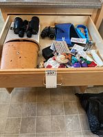 Full drawer view showing large vintage binocular with leather case, two smaller binoculars, magnets, and kitchen tools scattered inside wooden drawer