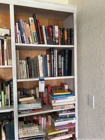 Right side of large bookshelf showing six shelves filled with books; bookshelf frame is white wooden with natural wood back panel; titles and authors of various books are visible in image.