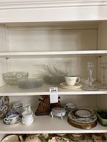 Shelf view showing clear glass plates, clear glass bowl, milk glass creamer and sugar set, cups, saucers, decorative plates, wooden bookends, and glass candlesticks.