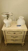 Two cream-colored vintage-style nightstands each with three drawers and ornate metal handles holding two white hobnail milk glass lamps with metal bases and glass chimneys.