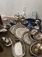 Wide angle of many silver and silver-plated items spread on floor including trays, bowels, teapots, cups, and serving dishes.