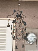 Large metal cat-shaped wind chime with decorative features and hanging clappers shown against wall with meter.