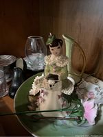 Shelf with various ceramic and porcelain figurines including a lady figurine in green dress, small toy shoe, pink flower pin, and clear glass item.