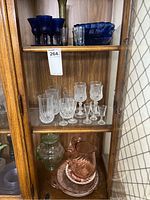 Full view of wooden cabinet shelf containing various blue, clear, pink, and green glassware pieces including goblets, pitchers, and plates.