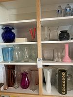 Shelves displaying multiple vases and glassware of different colors and shapes including blue, pink, clear, amber, purple, white, and black with floral decoration.
