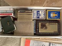 Top view of box containing multiple decks of playing cards including Bee brand and Hershey's branded tin along with other card boxes and assorted booklets.