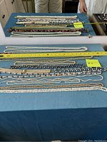 Twelve necklaces laid on blue cloth, different bead sizes and colors including white and dark beads, measuring tapes for length shown.