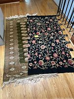 Both wall hangings laid flat on floor showing full size and pattern; black embroidered hanging on right with green wool rug on left