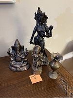 Three metal statues and one small vase on wooden surface with measuring tape for height reference.