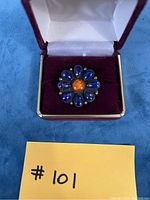 Front view of Ray Begay brooch with blue teardrop-shaped stones arranged in a flower pattern and a central orange round stone, set in silver with scalloped edges, included in a velvet display box.