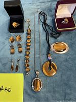 Image showing 4 pairs of tiger's eye earrings, 2 rings, a brooch, 2 necklaces, and a bracelet laid out on blue cloth; measuring tape included for scale.