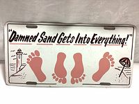 Front of metal sign features the text "Damned Sand Gets Into Everything!" with red footprints, lighthouse, and umbrella graphics on white metal.
