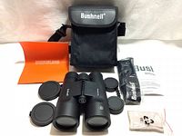 Bushnell binoculars with lens caps, carrying case, quick start guide, neck strap, and cleaning cloth displayed on surface.
